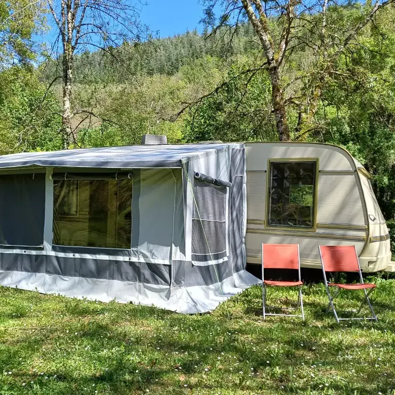 Large family caravan with a full-length awning attached, featuring large windows and two orange folding chairs set on green grass in a secluded, wooded spot.